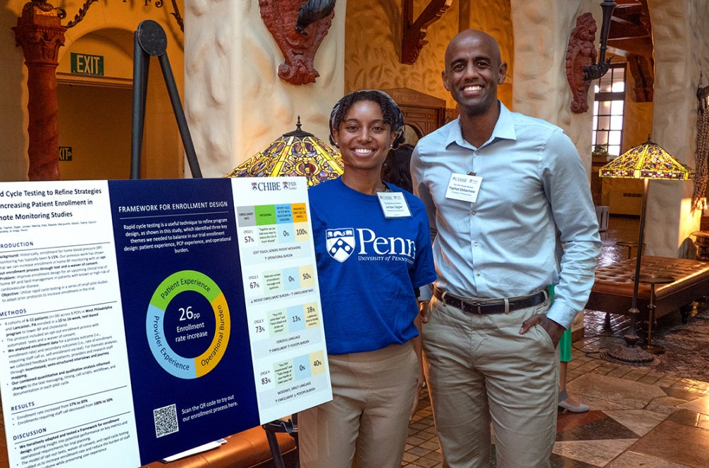 Woman in Penn logo t-shirt and a man posing next to a trifold presentation mounted on an easel