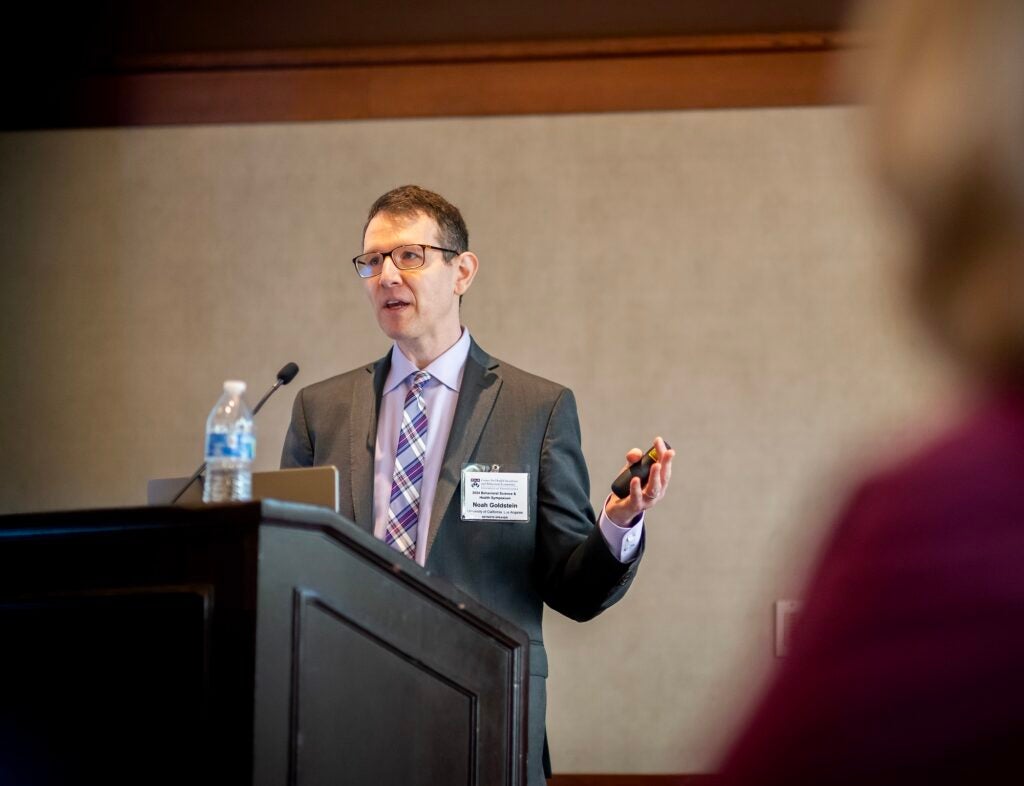 Noah Goldstein, in front of a podium, addresses symposium attendees. 